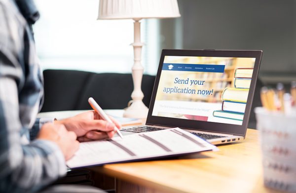 Young man writing college or university application form with pen and paper. Student applying or filling document. Scholarship, grant or admission. School website in laptop.